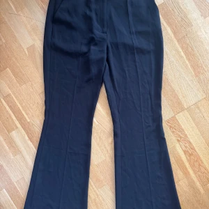 Svarta kostymbyxor från NA-KD - Snygga svarta kostymbyxor från NA-KD i klassisk modell med utsvängda ben och hög midja. Byxorna har bälteshällor, sidofickor och fuskbakfickor. Materialet är mjukt och har en dressad känsla, perfekt för en stilren look. 100% polyester. Namn: Flared Tailored Suit Pants - NA-KD. Nästintill oanvända 