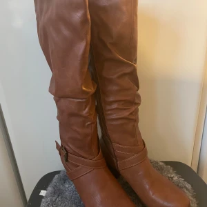 Bruna stövlar med klack  - Snygga bruna stövlar med dekorativt spänne och dragkedja på sidan. Stövlarna har en låg klack och är fodrade med mjukt fuskpälsfoder för extra värme.  med rund tå och platt sula framtill. Endast provade, stl 39