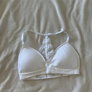 Vit ribbad bh med spetsdetalj - Supersöt vit bh med ribbad struktur och dekorativ spets i ryggen. Axelbandslös design med justerbart band upptill. Perfekt för att ge en extra touch till din outfit. Spetsen har ett blommigt mönster som gör toppen extra feminin och unik.