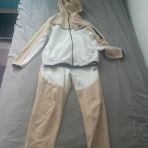 Nike mjukisbyxor beige och vita - Nike mjukisbyxor i beige och vitt med svart Nike-logga på höften. Byxorna har snörning i midjan och är gjorda i mjukt syntetmaterial. De har en relaxed passform och passar perfekt för chill eller träning. Snygg färgblockad design som sticker ut.