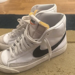 Nike Blazer Mid vita och svarta sneakers - Nike Blazer Mid sneakers i vitt skinn med svart swoosh och klassisk hög siluett. Skorna har snörning, vit sula och retroinspirerad design med synlig skumkant på plösen. Perfekta för dig som gillar en clean och tidlös look.