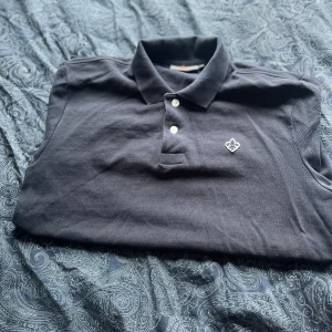 Mörkblå pikétröja,Morris - Säljer en mörkblå pikétröja från Morris med klassisk krage, två vita knappar och snygg broderad logga på bröstet. Tröjan är i bomull och har korta ärmar, perfekt för en stilren och avslappnad look.