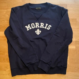 Mörkblå sweatshirt från Morris - Snygg mörkblå sweatshirt från Morris med vit broderad logga och emblem på bröstet. Sitter i normal passform och är perfekt till vintern. 9/10 skick. Hör av er för fler frågor.                   (Fraktar inom 24h)