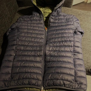 Mörkblå dunjacka från save the duck  - Säljer en snygg mörkblå dunjacka från save the duck och orange detaljer på dragkedjan. Jackan är quiltad och har en normal passform, perfekt för kyliga dagar. Insidan är olivgrön och jackan har långa ärmar samt tvåvägsdragkedja framtill.