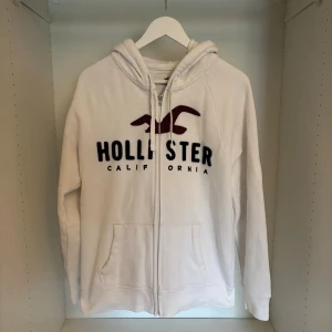 Vit hoodie från Hollister  - Säljer en fet och trendig vit hoodie från Hollister med broderad logga och text i vinrött och svart på bröstet. Storlek M och perfekt skick. Har du funderingar är det bara att fråga på.