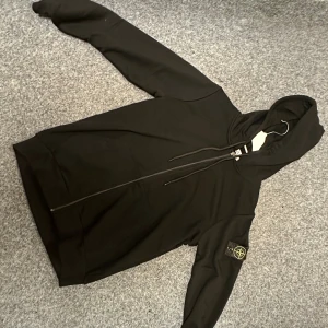 Svart hoodie från Stone Island - Svart hoodie från Stone Island med klassisk logopatch på vänster ärm. Tröjan har huva med snörning, hel dragkedja framtill och två fickor. Materialet är mjuk bomull och passformen är normal. Perfekt för dig som gillar streetwear och snygga detaljer.