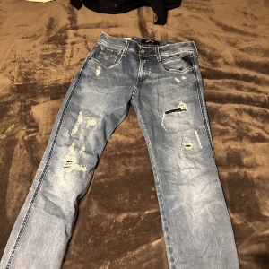 Replay Anbass slitna blå jeans, stl 29 - Säljer ett par Replay Anbass jeans i ljusblå tvätt med slitningar och hål för en riktigt edgy look. Slim fit-modell med klassiska fem fickor och snygga detaljer. Perfekta för dig som gillar streetstyle och vill sticka ut.