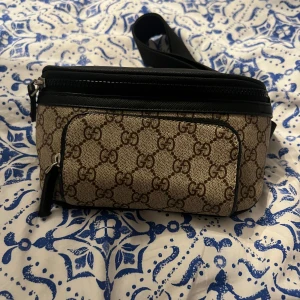  beige midjeväska med GG-mönster - Säljer en snygg beige midjeväska med klassiskt GG-mönster i brunt. Väskan har svarta detaljer, justerbart band och flera dragkedjefack. Perfekt för att bära det viktigaste och ge din outfit en lyxig touch.