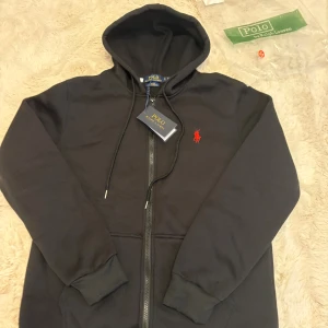 Svart hoodie från Polo Ralph Lauren - Svart hoodie från Polo Ralph Lauren med röd broderad logga på bröstet. Slim fit-modell med huva och snörning, dragkedja framtill och ribbade muddar. Tillverkad i mjuk bomull med stretch för extra komfort. Perfekt för chill dagar och streetwear.