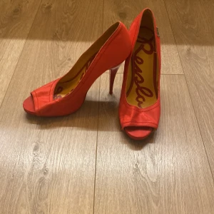 Röda pumps med öppen tå från Replay - Snygga röda pumps från Replay med öppen tå och hög klack. Skorna har en rundad form framtill och är tillverkade i ett tygmaterial med en lätt glansig finish. Insidan har Replay-logga i kontrastfärg. Perfekta för dig som vill sticka ut med färg.