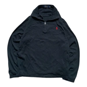 Svart half zip tröja från Polo Ralph Lauren - Klassisk svart långärmad tröja från Polo Ralph Lauren med röd broderad logga på bröstet. Tröjan har half zip-dragkedja vid halsen och är tillverkad i 100% pima bomull för extra mjuk känsla. Perfekt för lager-på-lager under höst och vinter. Har ett litet hål som syns på sista bilden
