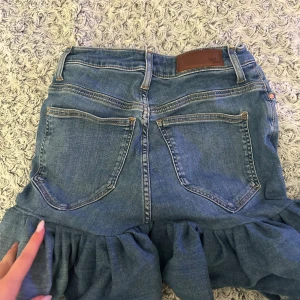 Jeanskjol med volang från Wrangler - Säljer en blå jeanskjol från Wrangler med volang nertill. Kjolen har klassiska jeansfickor både fram och bak, bälteshällor och knappstängning. Snygg och trendig modell som ger en cool vibe till outfiten.