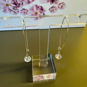 Silvriga örhängen med Swarovski kristaller  - Stilrena örhängen i silverfärgad metall med lång, droppformad båge och hängande Swarovski kristaller i 4mm som skimrar i regnbågens färger. Perfekta för dig som gillar minimalistisk design med en glittrig touch.