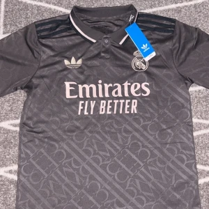 Mörkgrå Real Madrid fotbollströja - Snygg mörkgrå Real Madrid fotbollströja från Adidas med diskret mönster och broderade detaljer. Tröjan har korta ärmar, krage med knapp och klassiska Adidas-ränder på axlarna. Materialet är lätt och andas, perfekt för match eller träning. Size S. Fraktar inom 24 timmar.