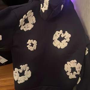 Mörkblå hoodie Denim Tears med vita blommor - Säljer en mörkblå hoodie från Denim Tears i storlek M står S men passar som M. Tröjan har huva och stora vita blommor på både fram- och baksida samt ficka framtill. Materialet är mjukt och insidan är fleecefodrad för extra komfort. Perfekt för dig som gillar streetwear och unika detaljer.