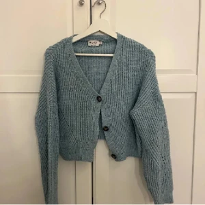 Ljusblå stickad kofta från NA-KD - Supermysig ljusblå stickad kofta från NA-KD med grov ribbad struktur och tre mörka knappar framtill. Koftan är croppad med lång ärm och v-ringad hals. Perfekt att slänga över en t-shirt för en chill look.