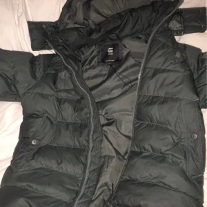 Mörkgrön pufferjacka från G-Star RAW - Säljer en mörkgrön pufferjacka från G-Star RAW med huva och dragkedja. Jackan är vadderad och har två fickor med knappstängning framtill. Perfekt för kalla dagar och har en riktigt schysst streetstil.