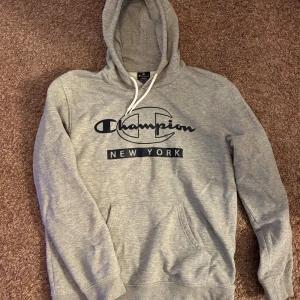 Grå hoodie från Champion New York - Snygg grå hoodie från Champion med stor svart logga och texten 'New York' på bröstet. Tröjan har huva med vita snören och en klassisk känguruficka framtill. Perfekt för en chill och sportig stil.