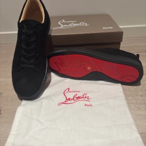 Svarta sneakers från Christian Louboutin - Snygga svarta sneakers från Christian Louboutin med klassisk röd sula och rund tå. Skorna har snörning och är tillverkade i mocka med mjukt beige foder. Perfekt för dig som vill ha en exklusiv och stilren look med en ikonisk detalj under foten.