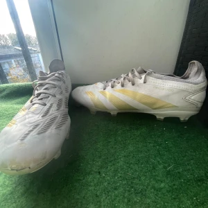 Adidas Predator day spark Fg pro  - Säljer ett par Adidas Predator fotbollsskor i vitt med guldiga detaljer och klassiska ränder. Skorna har snörning och platt sula med dobbar för gräsplan. Snygg, sportig design med rund tå och mönstrade paneler på ovansidan. Perfekt för dig som vill sticka ut på planen.