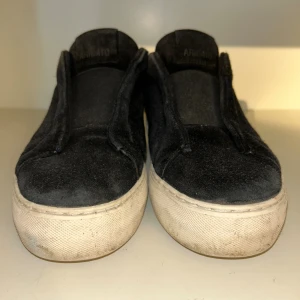 Svarta sneakers från Axel Arigato - Svarta sneakers från Axel Arigato med ovandel i mjuk mocka och vit platt sula. Minimalistisk design utan snörning, med rund tå och diskret branding på plösen. Insidan är fodrad med svart skinn för extra komfort. Perfekt för dig som gillar stilrena och cleana sneakers.