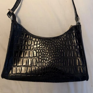 Svart axelväska  - Snygg svart axelväska med krokodilpräglat mönster och justerbar rem. Väskan har en stilren form och stängs med dragkedja upptill. Helt ny, aldrig använd.