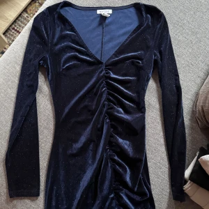 Mörkblå glittrig sammetsklänning NLY One - Supersnygg mörkblå sammetsklänning från NLY One med glittriga detaljer. Klänningen har lång ärm, v-ringning och snygg rynkning framtill som ger en cool slits. Perfekt för dig som vill sticka ut lite extra!