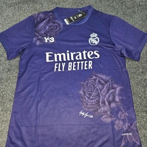 Real Madrid x Y-3 lila fotbollströja - Snygg lila Real Madrid fotbollströja från Adidas och Y-3 med blommigt rosmönster, klubbmärke och Emirates-tryck på bröstet. Tillverkad i lätt och ventilerande Aeroready-material, perfekt för match eller träning. Kortärmad och med modern design.