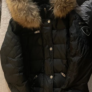 Svart dunjacka Rockandblue med äkta päls - Snygg svart dunjacka från Rockandblue med stor huva och äkta päls. Jackan har quiltade detaljer, dragkedjor på fickorna och silvriga knappar framtill. Perfekt för kalla dagar!