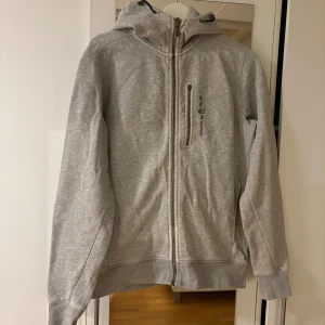 Grå hoodie från Sail Racing - Grå hoodie från Sail Racing med dragkedja framtill och en bröstficka med dragkedja. Tröjan har huva med snörning och logga på bröstet. Perfekt för dig som gillar en clean och sportig stil. Blixtlåsgrejen på bröstet är borta därav priset.