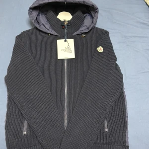 Moncler Cardigan Mörk blå - Säljer en mörkblå pufferjacka från Moncler med stickade ärmar och sidor, quiltad rygg och huva i glansigt material. Jackan har dragkedja framtill och två sidofickor med dragkedja. Moncler-logga på vänster ärm. Perfekt för dig som gillar snygga detaljer.