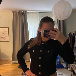 Svart croppad kofta med guldknappar - Snygg svart croppad kofta från H&M med dekorativa guldknappar framtill och på bröstfickorna. Koftan har lång ärm och är tillverkad i ett mjukt, stickat material som ger en cool och stilren look. Perfekt att styla med höga jeans eller kjol.