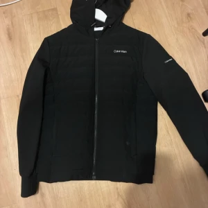 Calvin Klein - Svart pufferjacka från Calvin Klein med huva och dragkedja framtill. Jackan har diskret logga på bröstet och ärmen, quiltad framsida och släta ärmar. Perfekt för kyligare dagar.