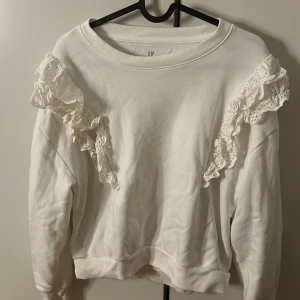 Vit sweatshirt med spetsvolanger GAP - Supersöt vit sweatshirt från GAP med snygga spetsvolanger på axlarna. Tröjan har rund hals, långa ärmar och är gjord i mjuk bomull. Perfekt för dig som gillar en clean look med lite extra detaljer. Volangerna ger en unik och trendig vibe.