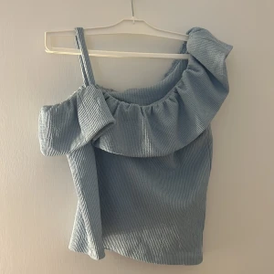 Ljusblå offshoulder topp med volang - Superfin ljusblå topp i ribbad manchester med en axelbandslös design och bred volang runt halsen. Toppen har en axelrem och en ärm, vilket ger en cool oneshoulder-look. Perfekt för dig som vill sticka ut med en trendig och unik stil.