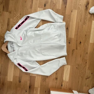 Vit Nike hoodie med rosa detaljer - Nike hoodie i vitt med dragkedja, huva och långa ärmar. Snygga rosa och svarta detaljer längs ärmarna med Nike-loggor och en rosa swoosh på bröstet. Tillverkad i mjuk polyester, perfekt för en sportig look. Skriv om du har några funderingar 