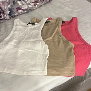 3-pack ribbade linnetoppar från Zara - Säljer tre ribbade linnetoppar från Zara i storlek M. Färgerna är vit, beige och rosa. Alla toppar är croppade och har en klassisk ärmlös design med rund halsringning. Perfekta att matcha med jeans eller kjol för en chill look.