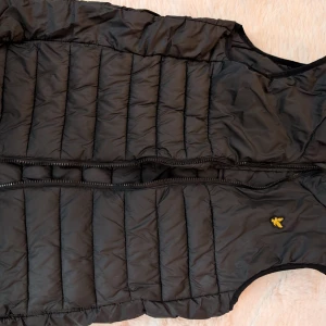 Svart dunväst från Lyle & Scott - Säljer en svart dunväst från Lyle & Scott med klassisk gul logga på bröstet. Västen har quiltad design, dragkedja framtill och är perfekt att slänga över en hoodie eller tröja. Enkel och stilren look som funkar året runt.Kan även användas under en jacka som extra lager. Strl 12-13 år. 
