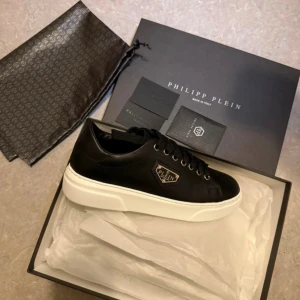 Svarta sneakers från Philipp Plein - Snygga svarta sneakers från Philipp Plein med vit platt sula och metallogo på sidan. Skorna har snörning och är tillverkade i skinn. Perfekta för dig som gillar exklusiv streetstyle och vill sticka ut med en lyxig detalj. Använd 1 gång 