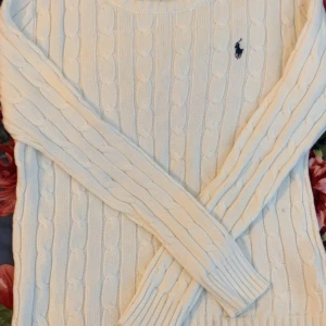 Vit kabelstickad tröja Polo Ralph Lauren - Kabelstickad vit tröja från Polo Ralph Lauren med klassisk marinblå logga broderad på bröstet. Tröjan har rund halsringning, långa ärmar och ribbade muddar. Perfekt för lager-på-lager och snygg till jeans eller kjol.