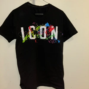 Svart ICON t-shirt med färgstänk - Svart t-shirt med stort vitt 'ICON'-tryck framtill och coola färgstänk i grönt, blått, rött och rosa. Klassisk rund hals och korta ärmar. Perfekt för dig som vill sticka ut med en streetstyle-look.