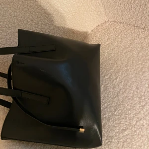 Svart axelväska i skinn med gulddetalj - Stilren svart axelväska i skinn med minimalistisk design och raka linjer. Väskan har långa handtag och en liten gulddetalj på framsidan. Perfekt storlek för att få plats med allt du behöver och passar till många olika outfits.