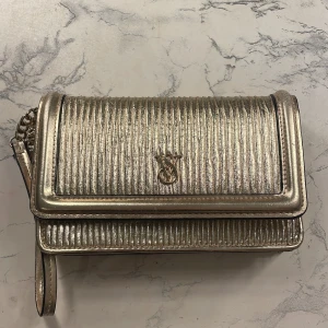 Guldig handväska från Victoria's Secret - Snygg guldig handväska från Victoria's Secret med metallic finish och vertikala ränder. Plånboken har ett stilrent VS-emblem i metall, kedjedetalj och flera fack för kort. Materialet är skinnimitation med glansig yta och praktisk knappstängning.✨🩷