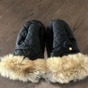 Svarta quiltade vantar med fuskpäls - Snygga svarta quiltade tumvantar från BOSS med mjuk beige fuskpäls runt handleden. Vantarna har ett elegant rutmönster och en metallbricka med logga. Perfekta för kalla vinterdagar när du vill hålla stilen.