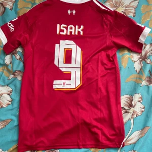 Liverpool röd fotbollströja Adidas S - Liverpool fotbollströja från Adidas i röd färg med vita detaljer och tryck. Nummer 9 och namnet ISAK på ryggen, samt UEFA Foundation-märke på ärmen. Slim fit-modell i lätt och ventilerande polyester. Klassiska Adidas-ränder på axlarna och rund hals.