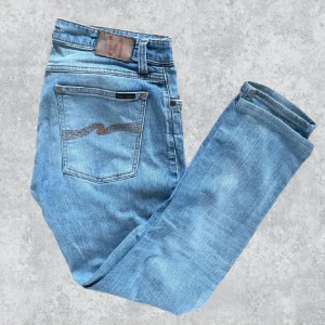 Nudie - Säljer ett par ljusblå jeans från Nudie Jeans med klassisk femficksdesign och snygga kontrastsömmar. Jeansen har avsmalnande ben och en skön passform, med Nudie-logga på bakfickan och läderpatch i midjan. Perfekt för dig som gillar stilrena och tidlösa jeans. Storlek W32 L30