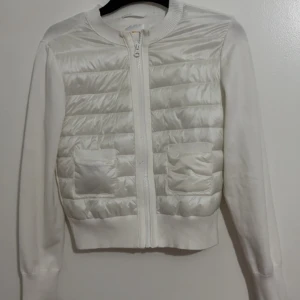 Vit quiltad kofta från Gina Tricot - Snygg vit kofta från Gina Tricot med quiltad framsida och två fickor. Dragkedja hela vägen och ribbade muddar. Ärmar och baksida i mjukt tyg, framsidan har glansig finish. Perfekt för lager-på-lager under kyliga dagar.