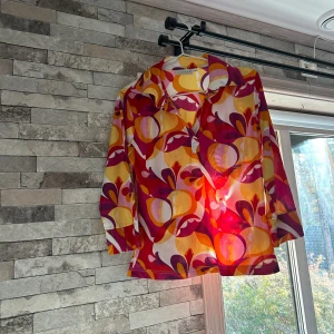 Färgglad retro skjorta från Hennes - Säljer en färgstark retro skjorta från Hennes Collection med galet snyggt mönster i rött, orange, gult, lila och vitt. Skjortan har lång ärm, klassisk krage och knäppning framtill. Perfekt för dig som vill sticka ut med 70-talsvibbar!