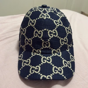 Mörkblå Gucci keps  - Säljer en mörkblå keps från Gucci med ikoniskt GG-mönster i beige över hela kepsen. Kepsen har justerbar rem baktill i läder med präglad Gucci-logga. Materialet är strukturerat och ger en lyxig känsla. Perfekt accessoar för att lyfta din outfit.