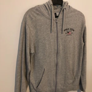 Grå hoodie från Polo Club med dragkedja - Grå hoodie från Polo Club Saint Vincent med tryck på bröstet. Tröjan har huva med snörning, hel dragkedja framtill och två fickor. Mjuk bomullskänsla och långärmad modell, perfekt för chill dagar eller när du vill vara extra comfy.